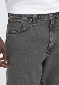 Blugi din denim gri cu croială clasică, prevăzuți cu buzunar frontal, cusături subtile și accente din metal. Materialul are un finisaj texturat.