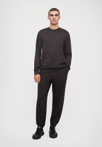Mørkebrun sweatshirt med rund hals og lange ermer, kombinert med matchende smale bukser og svarte joggesko, satt mot en ensfarget bakgrunn.