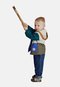 Enfant portant une veste multicolore avec des sections vertes, beiges et bleues, un jean bleu et des sandales à motifs, tenant une branche avec assurance.