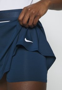 Tmavě modrá sportovní sukně s textura a bílým logem Nike, držená tak, aby odhalila odpovídající přiléhavé šortky pod ní.