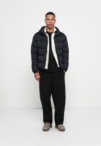 Puffer jacket in navy with a white zip-up inner layer, paired with black cargo pants and grey shoes. Smooth texture, relaxed fit, and ribbed cuffs.