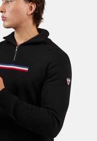 Schwarzer Reißverschluss-Pullover mit strukturiertem Strick, der ein horizontales Streifenmuster in Rot, Weiß und Blau aufweist. Logopatch am Ärmel.