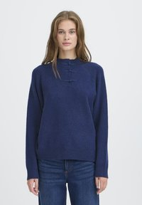 Jauna sieviete ar gariem gaiši brūniem matiem, tērpusies tumši zila džemperī ar trim dekoratīvām horizontālām cilpām un zilām džinsām, stāv pret baltu fona.