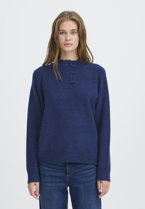 Young woman with long light brown hair wearing a navy blue sweater with three decorative horizontal loops and blue jeans, standing against white background.