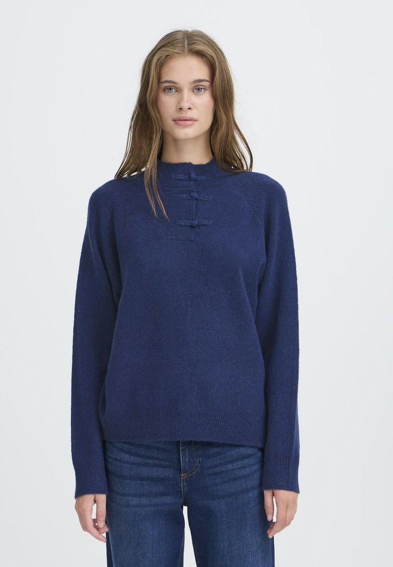 Jauna sieviete ar gariem gaiši brūniem matiem, tērpusies tumši zila džemperī ar trim dekoratīvām horizontālām cilpām un zilām džinsām, stāv pret baltu fona.