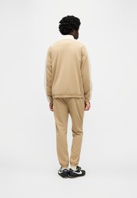 Μπεζ φόρμα με ζακέτα με φερμουάρ και ελαστικά μανίκια, με άσπρη λεπτομέρεια στους ώμους. Μαύρα αθλητικά παπούτσια με λευκές λεπτομέρειες.