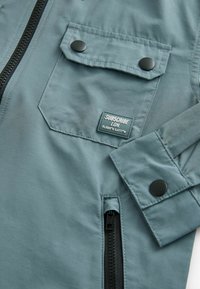 Giacca blu chiaro con texture opaca, dotata di chiusura con zip, due tasche frontali, un'etichetta con il logo "SUBSCRIBE LDN" e dettagli metallici neri.