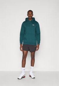 Felpa con cappuccio color teal, con tasca a marsupio e logo sul petto, abbinata a pantaloni corti scuri e sneakers bianche con accenti rossi.