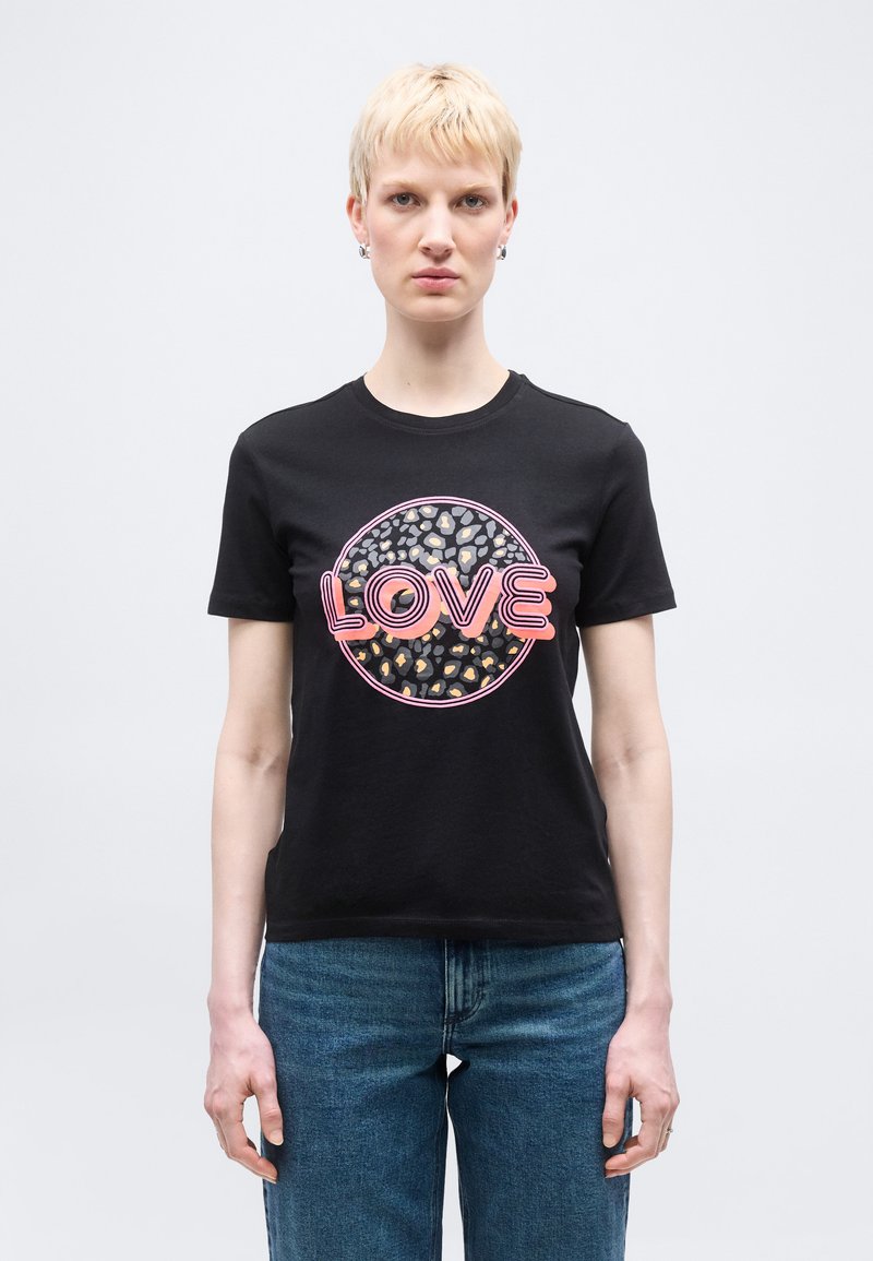 Άτομο με κοντά ξανθά μαλλιά που φοράει μαύρο μπλουζάκι με ροζ κείμενο «LOVE» και κύκλο με μοτίβο λεοπάρδαλης, συνδυασμένο με μπλε τζιν, στέκεται μπροστά σε λευκό φόντο.