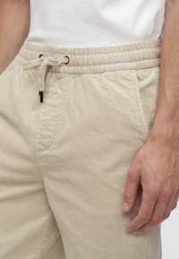 Close-up of a person wearing beige corduroy pants with an elastic waistband and drawstring, paired with a white shirt.
