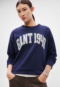 Tengerészkék pamut pulóver fehér "GANT 1949" nyomtatott felirattal, környakú kivágással, bordázott mandzsettákkal és laza szabással. A modell zsebbe dugott kézzel pózol.