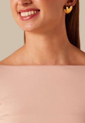 Smiling woman wearing gold heart-shaped earrings and a light pink off-shoulder top against a beige background.