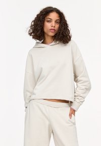 Lichtbeige cropped hoodie met een verstelbare capuchon, zachte textuur, oversized pasvorm en ruwe randdetails. Gecombineerd met bijpassende broek.