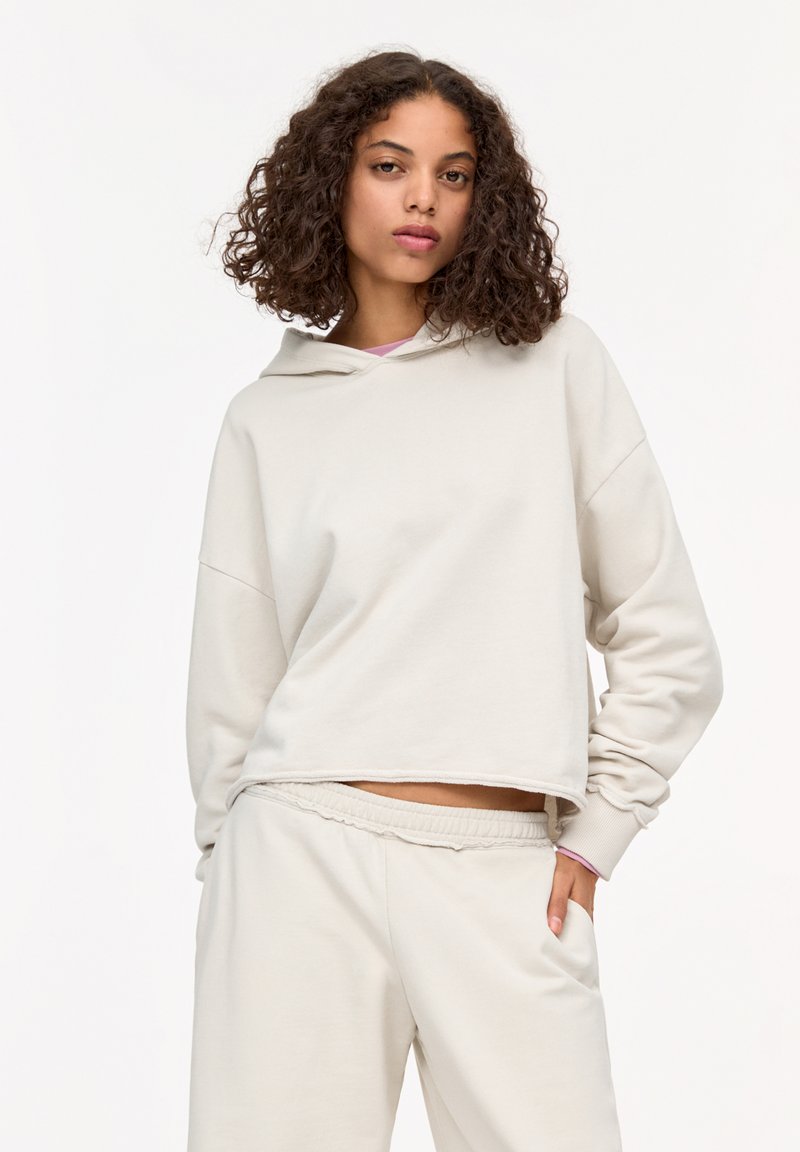Lichtbeige cropped hoodie met een verstelbare capuchon, zachte textuur, oversized pasvorm en ruwe randdetails. Gecombineerd met bijpassende broek.