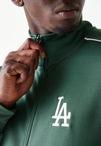 Πράσινο μπουφάν με φερμουάρ, κατασκευασμένο από λείο ύφασμα, με λευκό κεντημένο λογότυπο "LA" στο στήθος και λευκές λεπτομέρειες περιμετρικά.