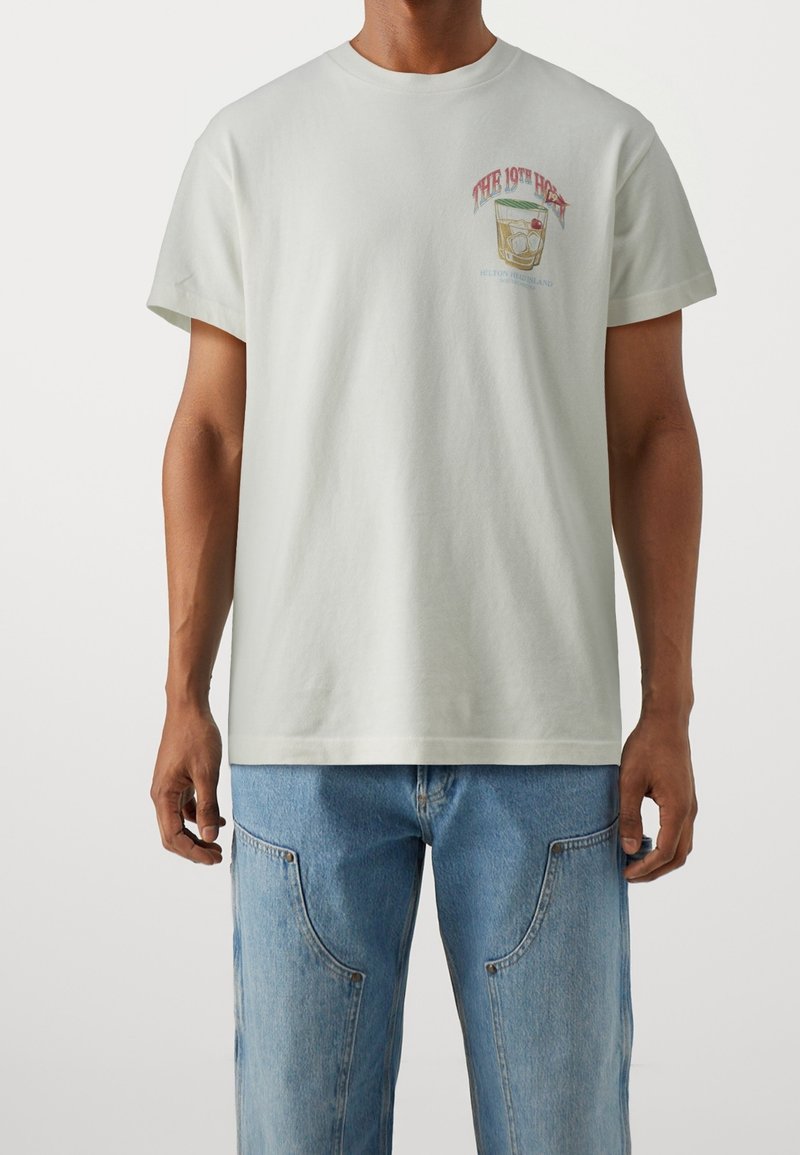 Ljusgrå t-shirt i bomull med kort ärm, med en färgglad grafisk tryck på bröstet och rund halsringning. Kombinerad med blå denimjeans.