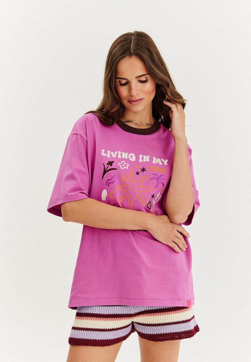 Růžové oversized tričko s krátkými rukávy, které má barevný grafický design a hnědý límec. Sestaveno s vícebarevnými háčkovanými šortkami.