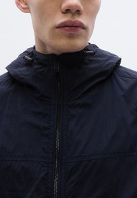 Tmavomodrá bundá s kapucňou na zips vpredu, vyrobená z ľahkej látky, s nastaviteľnými šnúrkami pri golieri. Hladký povrch, jednoduchý dizajn a detaily švov.