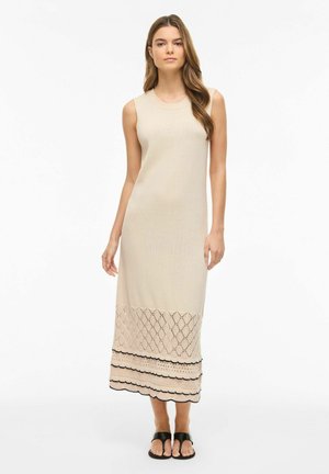 Kobieta stojąca w bezrękawnikowej beżowej dzianinowej sukni maxi z wzorzystym dołem i czarnych sandałach na białym tle.
