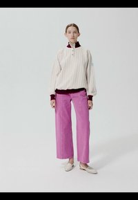 Donna in piedi che indossa un pullover crema a righe sottili con polsini e colletto bordeaux, pantaloni larghi rosa acceso e scarpe bianche su uno sfondo semplice.