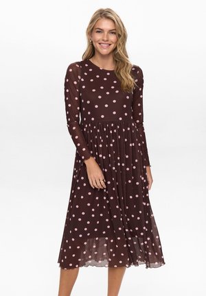 Brązowa sukienka z długim rękawem w różowe kropki, o luźnym fasonie, z okrągłym dekoltem i plisowaną spódnicą; materiał wydaje się lekki i prześwitujący.