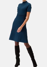 Stickad klänning i mörk teal med korta ärmar, ribbat mönster, vågig hög halsringning och utsvängd kjol. Tillsammans med svarta knähöga stövlar.