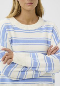 Femme aux bras croisés portant un pull en maille rayé blanc et bleu clair, visible des épaules au haut de la poitrine.
