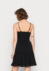 Vestido preto com alças finas, corpete franzido, bainha em camadas com ruffles e pequenos pontos texturizados. Tecido leve e silhueta justa.