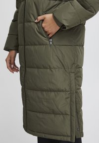 Garšs olīvu zaļš pūkains mētelis ar horizontālu quilted rakstu, sānu rāvējslēdzēja kabatām un elastīgiem aprocēm, izgatavots no gludas, ūdensnoturīgas vielas.