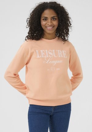 Peach-colored sweatshir t with white "LEISURE League" text. Ribbed collar and cuffs, soft fabric, relaxed fit, worn with blue jeans.