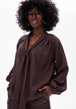 Bruine blouse met lange mouwen, voorzien van een strikdetail bij de nek en een subtiele textuur, gecombineerd met hoge taillebroeken, gepresenteerd op een model.