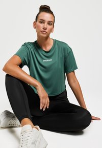 Haut de sport vert à manches courtes avec texture côtelée et encolure ronde, arborant un logo "Redmax" blanc, associé à des leggings noirs et des baskets.