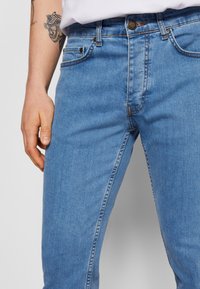 Világoskék farmernadrág egyenes szabású, öt zsebes dizájnnal rendelkezik, kontrasztos varrással és fém gombos záródással a deréknál.