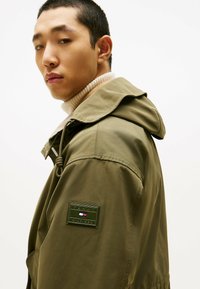 Lekka kurtka w kolorze oliwkowej zieleni z kapturem, z teksturowanym kołnierzem i naszywką na lewym rękawie z logo marki i jej kolorami.
