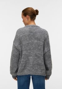 Γυναίκα με μαλλιά σε κότσο που φοράει ασημένια σκουλαρίκια, φαρδιά χνουδωτή γκρι πλεκτή μπλούζα και μπλε τζιν, στραμμένη προς τα πίσω με φόντο απλό.