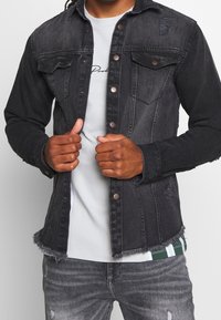 Chaqueta de mezclilla negra con un lavado desteñido, cierre de botones, bolsillos en el pecho y dobladillo deshilachado. Lleva puesta una camiseta gris claro con detalles de rayas verdes.