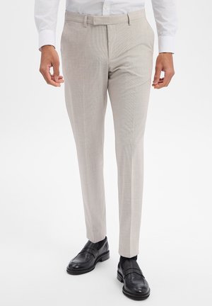 Man wearing slim-fit beige dress trousers with black leather loafers and a white dress shirt, standing against a plain white background.