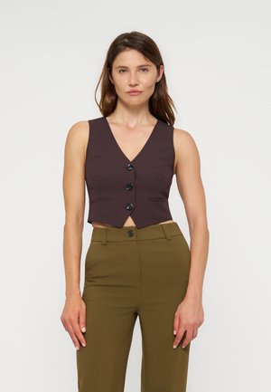 Femme portant un gilet sans manches marron foncé à boutons et un pantalon taille haute vert olive, debout sur un fond blanc uni.
