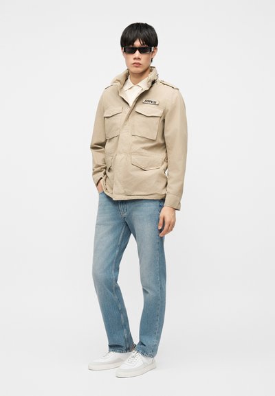 Homme portant des lunettes de soleil foncées, une veste beige avec des écussons, un jean bleu clair et des baskets blanches, debout avec une main dans la poche.