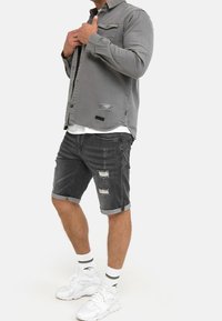 Grijs denimshirt met versleten plekken, gecombineerd met grijze denimshorts met opgerolde boorden. Draagt witte sneakers en gestreepte sokken.