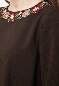 Närbild av en mörkbrun texturerad tygöverdel med broderad blommig halslinning som visar röda, rosa och krämfärgade blommor med gröna blad.