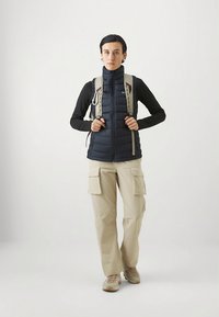 Granatowa puchowa kamizelka na czarnej koszulce z długim rękawem, beżowe spodnie typu cargo oraz jasne buty do lekkiego trekkingu; plecak z widocznymi paskami i elementami metalowymi.