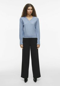 Hellblauer, strukturierter Strickpullover mit V-Ausschnitt und langen Ärmeln, kombiniert mit hoch taillierten schwarzen weit geschnittenen Hosen und spitzen schwarzen Schuhen.