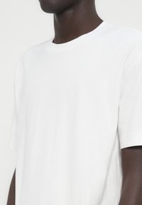 Κοντινό πλάνο ενός ατόμου που φοράει απλή λευκή κοντομάνικη μπλούζα, σε φόντο απλού λευκού χρώματος.