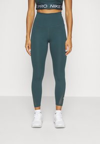 Legginsy w kolorze turkusowym z wysokim stanem, wykonane z elastycznego materiału z siateczkowymi akcentami przy kostkach i markowym pasem, w połączeniu z białymi trampkami.