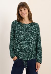 Grün gemusterte Bluse mit langen Ärmeln, rundem Ausschnitt und einem Bindeband am Saum. Verfügt über einen strukturierten Druck in dunkleren und helleren Farbtönen.