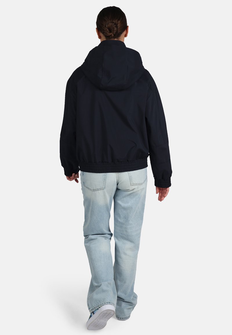 Granatowa kurtka z kapturem o luźnym kroju, z elastycznym pasem i mankietami, zestawiona z jasnoniebieskimi dżinsami. Materiał wydaje się gładki i trwały.