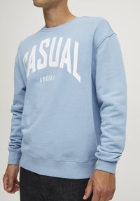 Svijetloplava dukserica od mekanog materijala, s odvažne bijele natpise "CASUAL FRIDAY" otisnute na prednjoj strani. Rebrasti rukavi i rub.