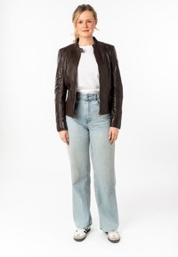 Giubbotto in pelle marrone con colletto alto, chiusura con zip e design aderente, abbinato a jeans a gamba larga blu chiaro e sneakers casual.