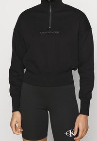 Sudadera negra con cremallera en el cuello, hecha de mezcla de algodón, con dobladillo y puños acanalados. Logotipo "CALVIN KLEIN JEANS" en el centro.